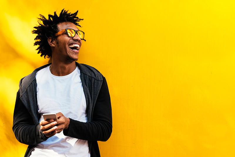 happy Black man wearing sunglasses using cell phone in front of bright yellow wall